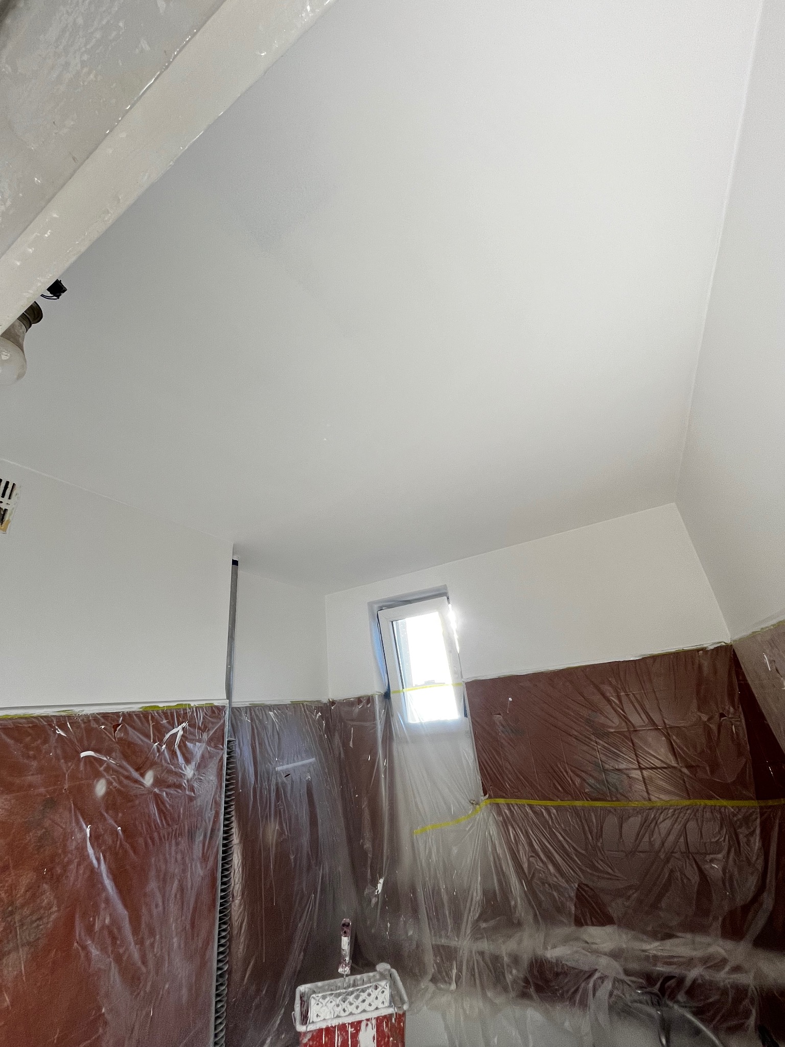 Białe ściany i sufit po malowaniu, zabezpieczone folią i taśmą malarską. Widoczny wałek i kuweta malarska. Jasne światło wpada przez okno.