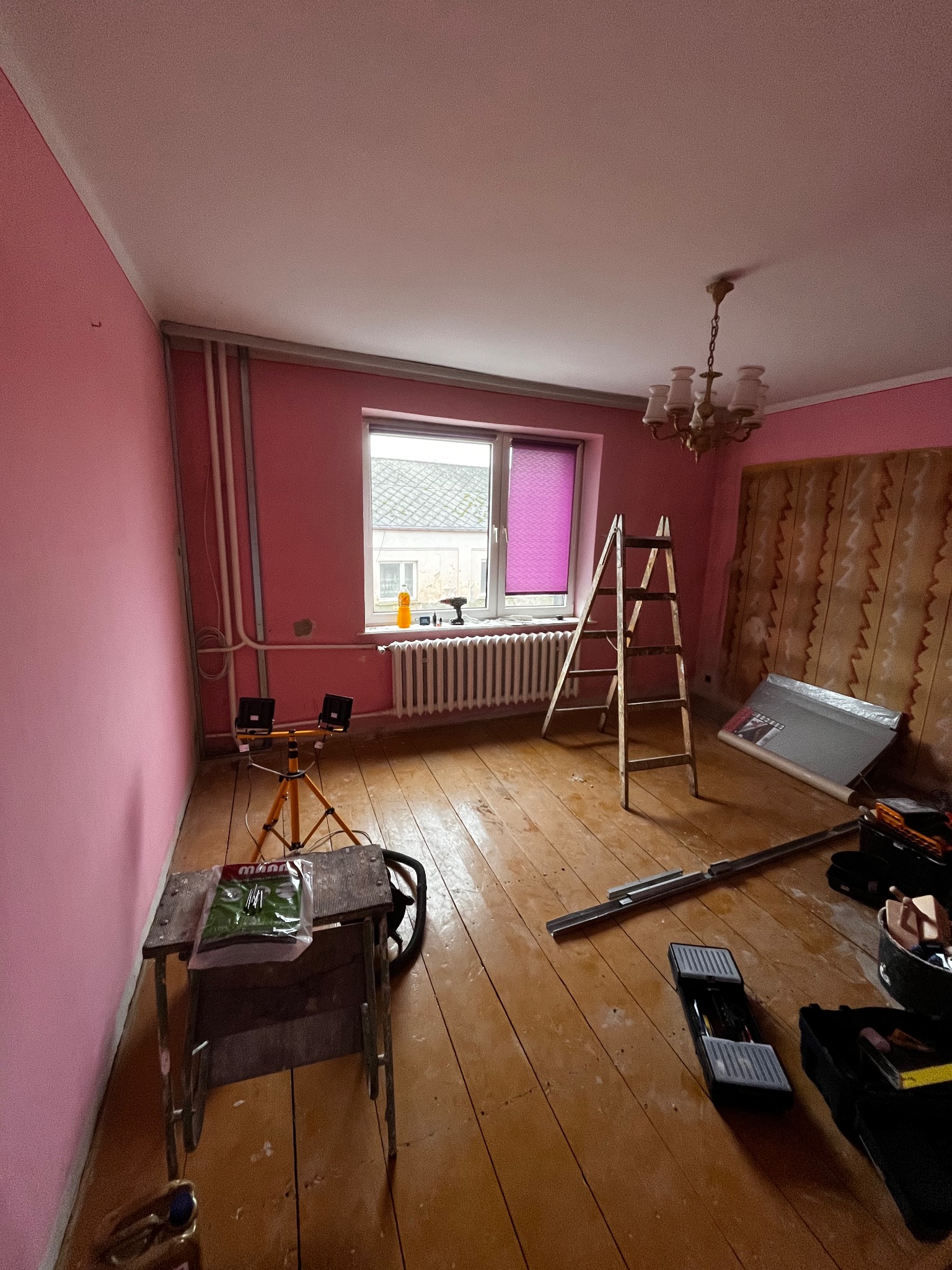 Pokój w trakcie remontu: różowe ściany, drewniana podłoga, drabina oparta o okno z fioletową roletą. Widoczne narzędzia i lampa na statywie. Częściowo zdjęta tapeta.