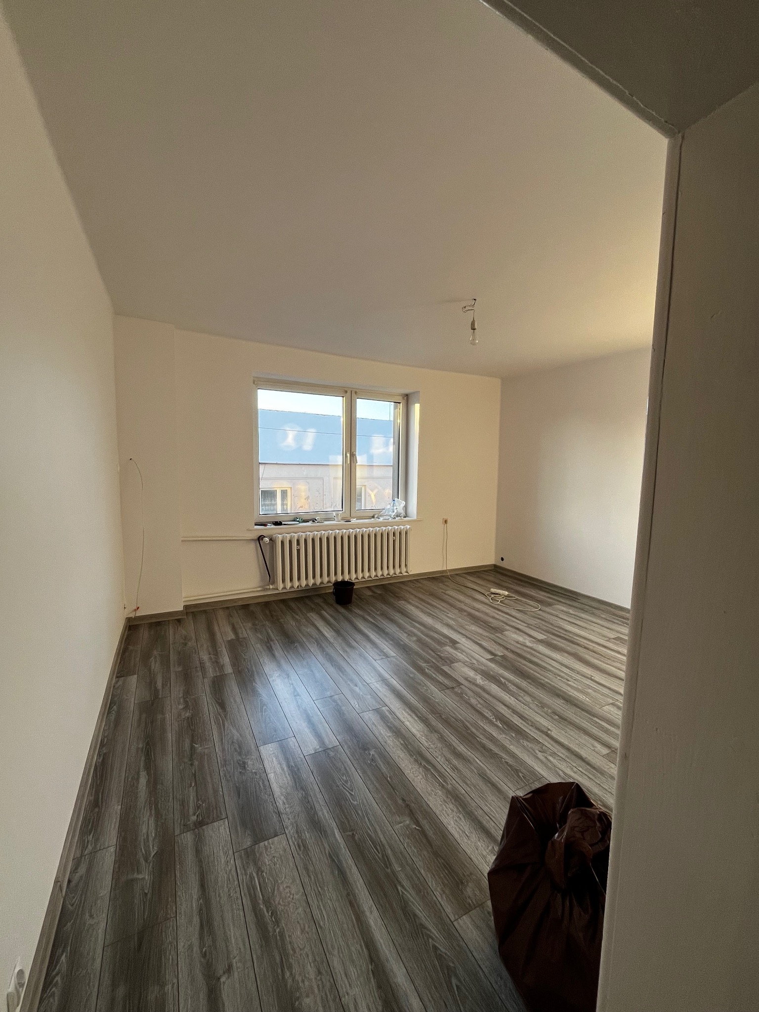 Puste, jasne wnętrze po remoncie: świeżo pomalowane ściany, ułożone panele podłogowe w odcieniach szarości, grzejnik pod oknem. Widoczny worek z odpadami.