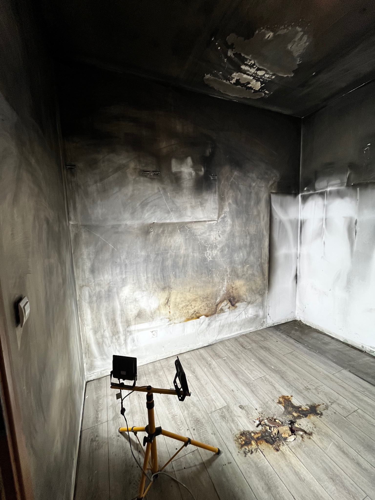 Pokój po pożarze: osmolone ściany, sufit i podłoga z widocznymi uszkodzeniami. Na podłodze lampa na statywie oświetlająca obszar zniszczeń.