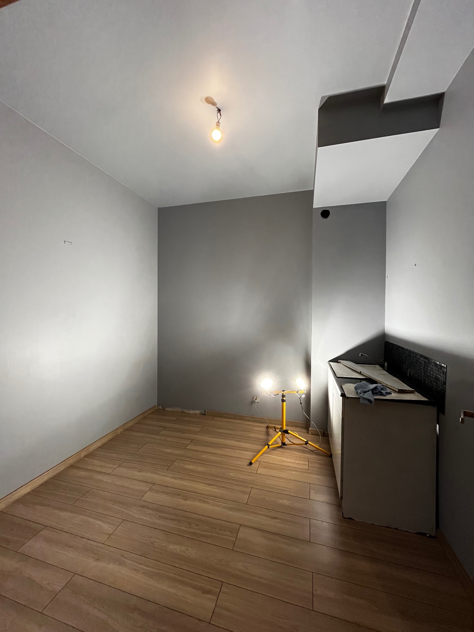 Szare wnętrze po remoncie: drewnopodobne płytki na podłodze, świeżo pomalowane ściany w odcieniach szarości, żółty statyw z lampami oświetlającymi przestrzeń.