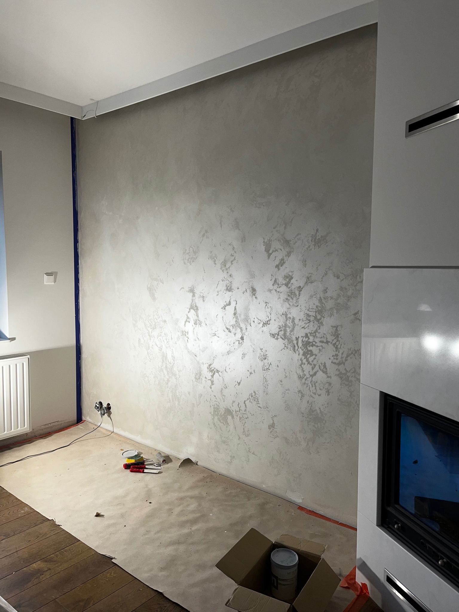 Ściana z efektem dekoracyjnym w odcieniach szarości, zabezpieczona podłoga, widoczne narzędzia malarskie i puszka farby. Fragment kominka z boku kadru.