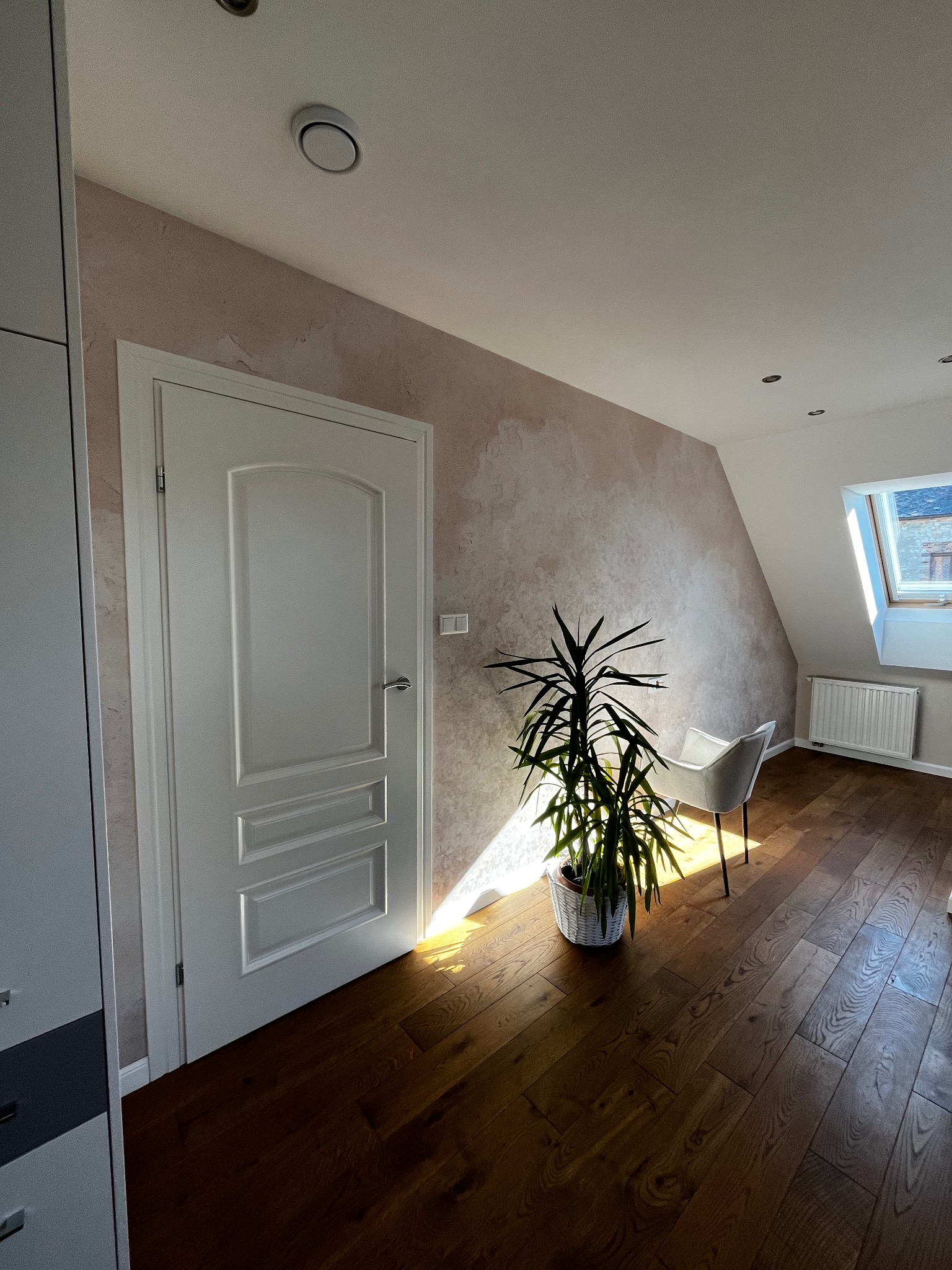 Jasne wnętrze poddasza z drewnianą podłogą, ścianą w odcieniach beżu i białymi drzwiami. W rogu stoi roślina doniczkowa, a przez okno wpada naturalne światło.