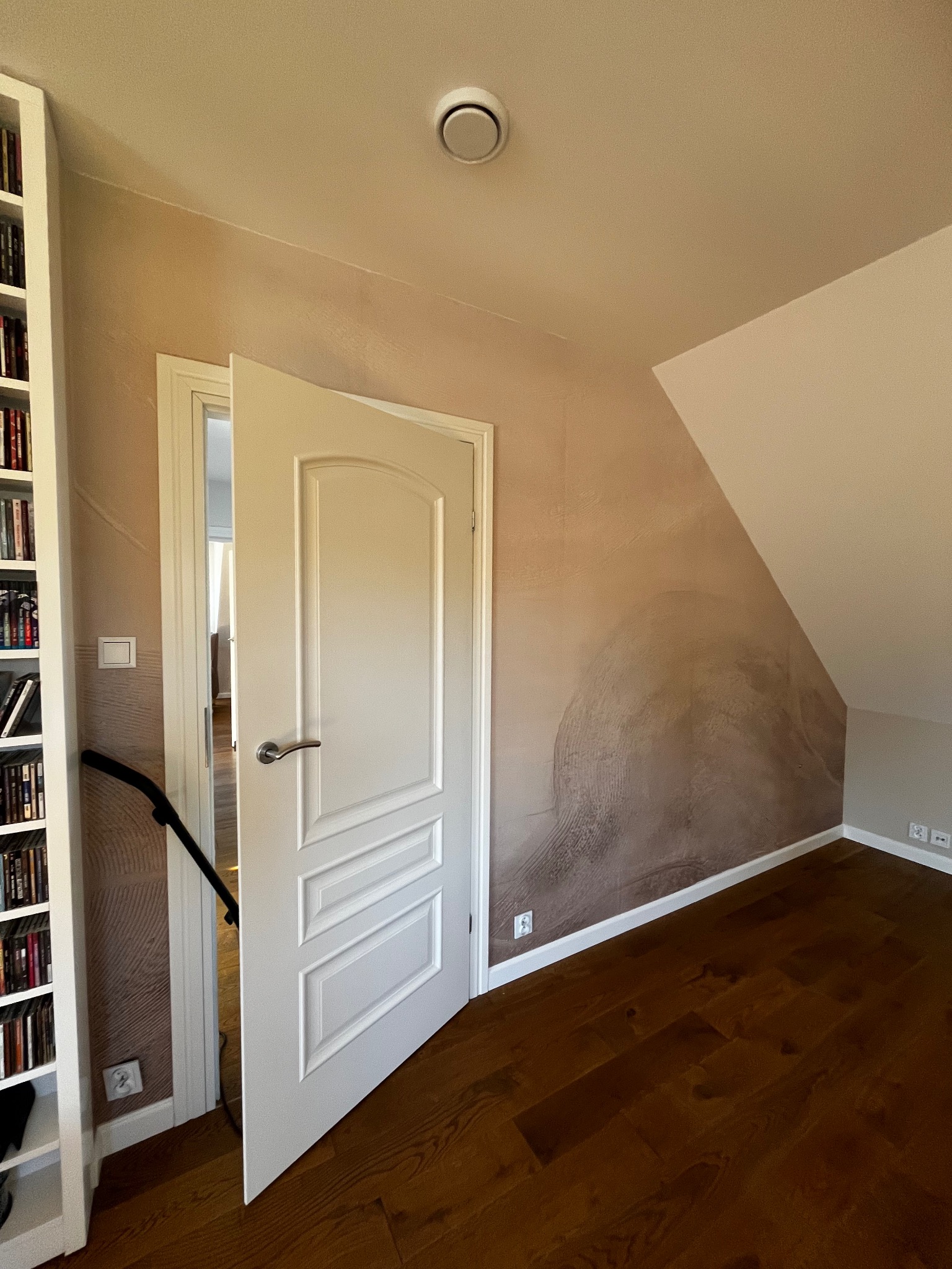 Pokój na poddaszu z drewnianą podłogą i ścianą z dekoracyjną, beżową fakturą. Otwarta biała, panelowa  drzwi. Widoczny fragment regału z książkami.