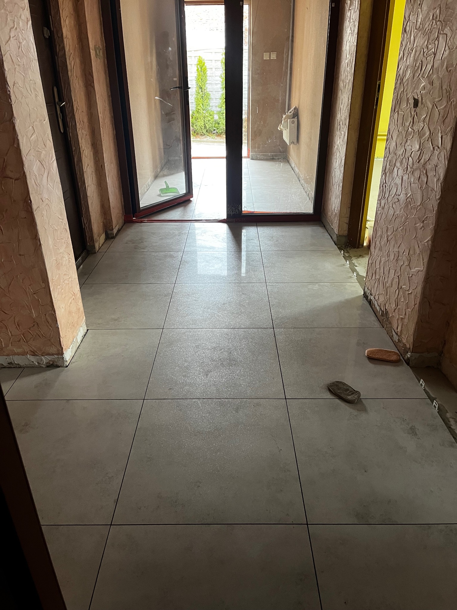 Ułożona jasnoszara podłoga z płytek w korytarzu z otwartymi drzwiami na zewnątrz. Widoczne narzędzia glazurnicze i surowe ściany z fakturą.