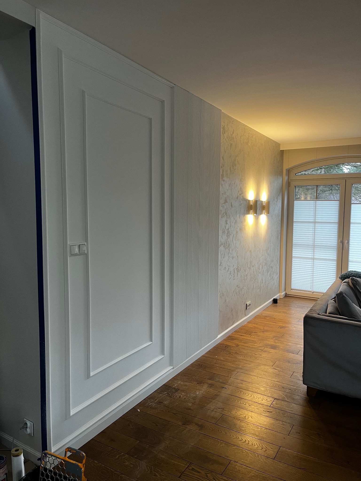 Eleganckie wykończenie wnętrza: białe panele ścienne z dekoracyjnymi listwami, wzorzysta tapeta i drewniana podłoga w ciepłym oświetleniu. Widoczny fragment sofy.