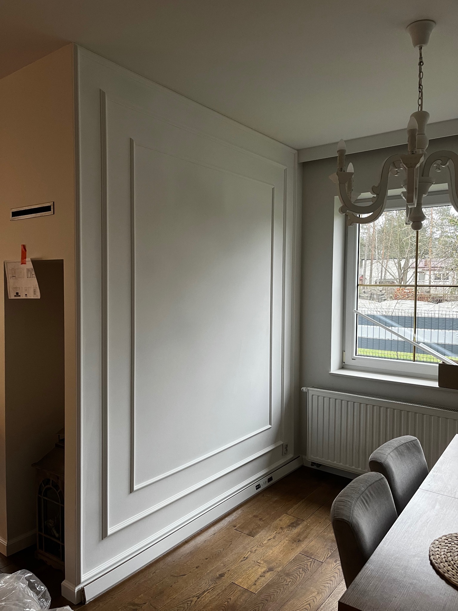 Elegancka ściana z dekoracyjnymi listwami w jasnym wnętrzu. Widoczny fragment parkietu, okno z widokiem na zewnątrz i ozdobny żyrandol. Subtelne wykończenie.