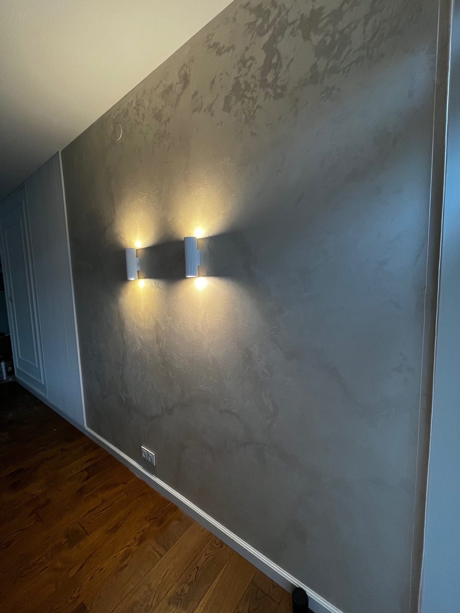 Ściana z efektem dekoracyjnym w odcieniach szarości, oświetlona dwoma nowoczesnymi kinkietami. Widoczny fragment drewnianej podłogi i gniazdko elektryczne.