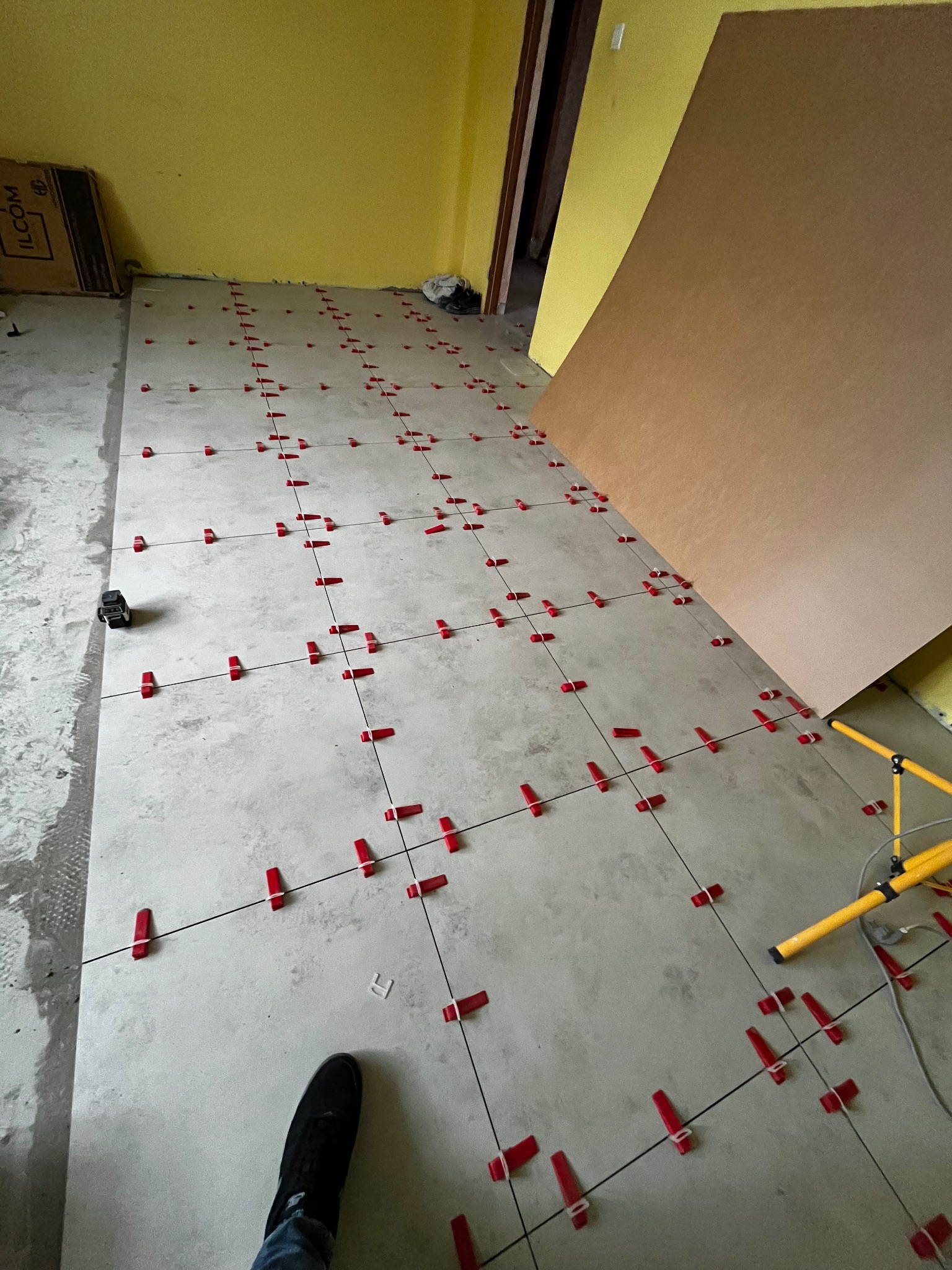 Układanie szarych płytek podłogowych z czerwonymi klipsami poziomującymi. Widoczny fragment buta i lampa oświetleniowa. Żółta ściana w tle.