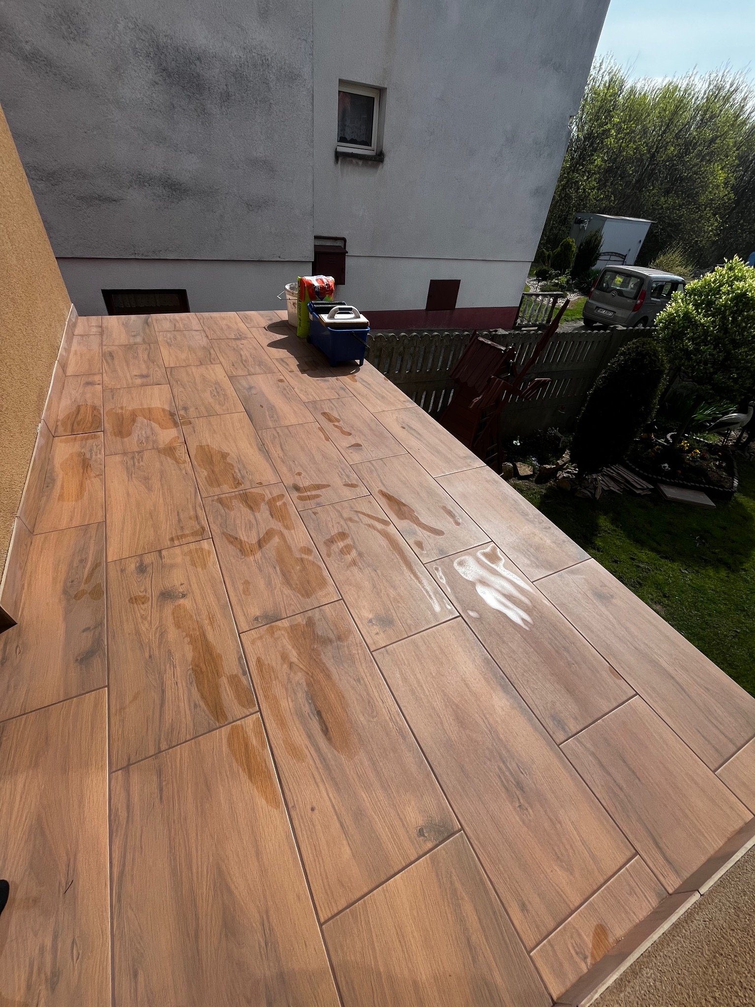 Balkon wyłożony płytkami imitującymi drewno, z widocznymi śladami wody, sugerujący świeżo zakończone prace glazurnicze. W tle budynek i zieleń.