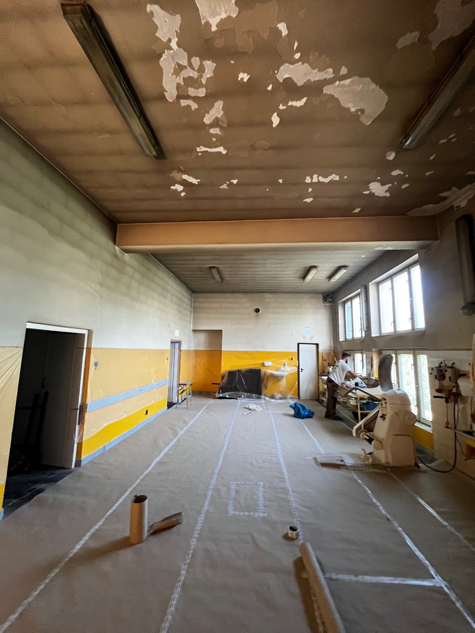 Przygotowanie do prac w dużej sali: zabezpieczona podłoga, ściany z żółtym pasem, widoczne odrapania na suficie. W tle pracownik przy maszynie.