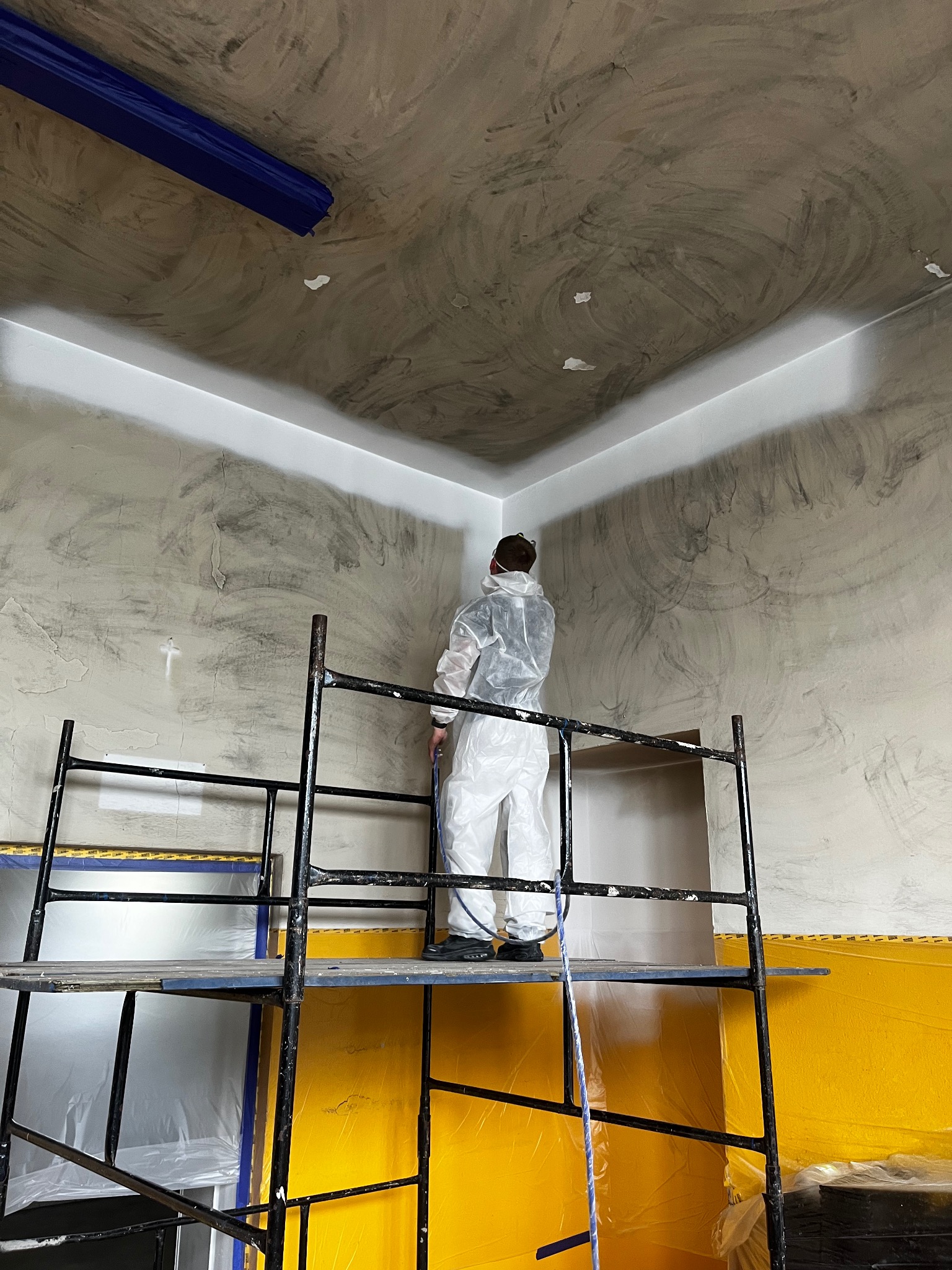 Pracownik w kombinezonie maluje natryskowo sufit i ściany z rusztowania. Widoczne zabezpieczenia folią i taśmą malarską. Ściany w odcieniach szarości, sufit częściowo biały.