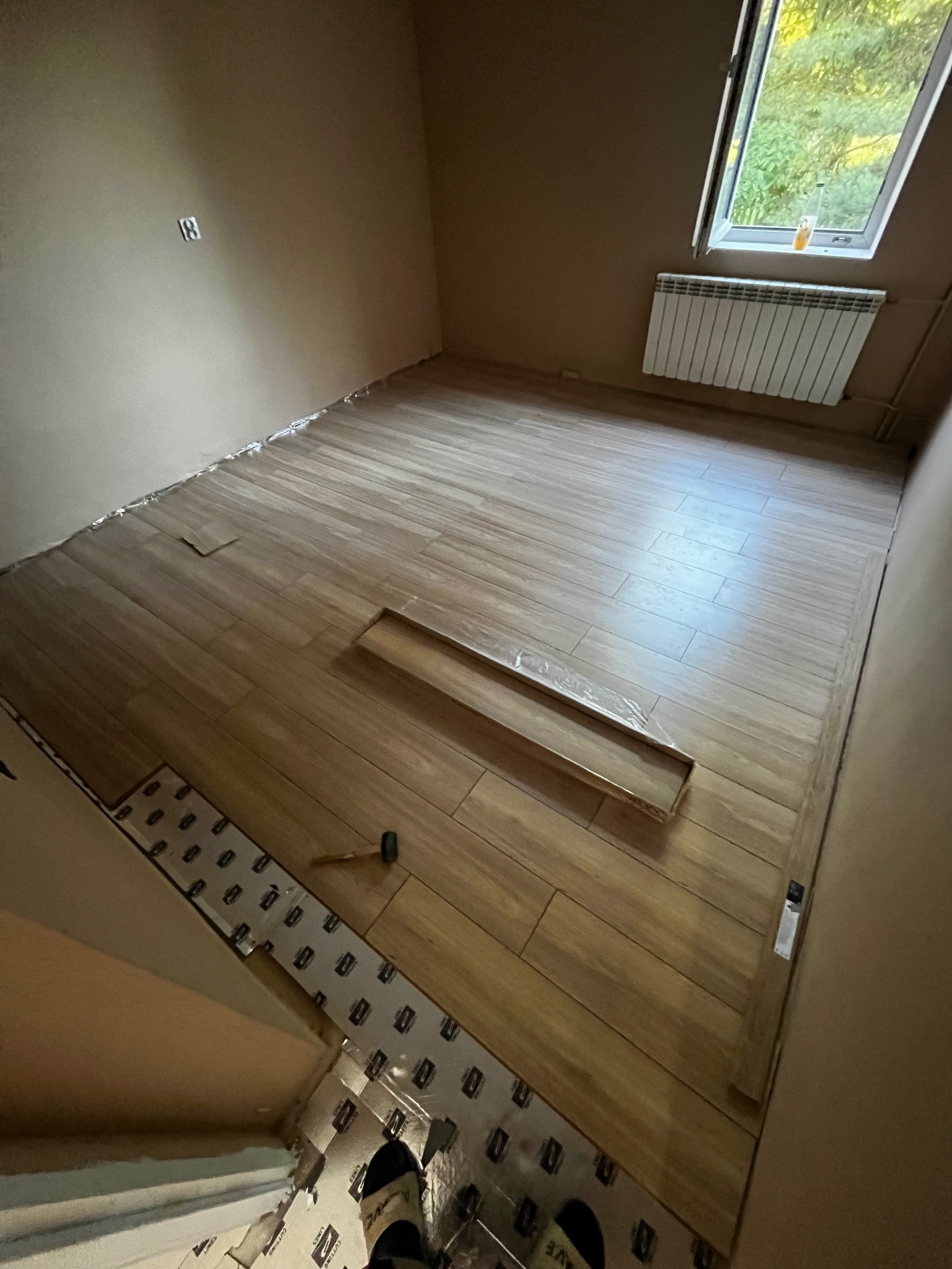 Układanie paneli podłogowych w pokoju z beżowymi ścianami. Częściowo ułożone panele, widoczna folia izolacyjna i młotek. Okno z grzejnikiem w tle.