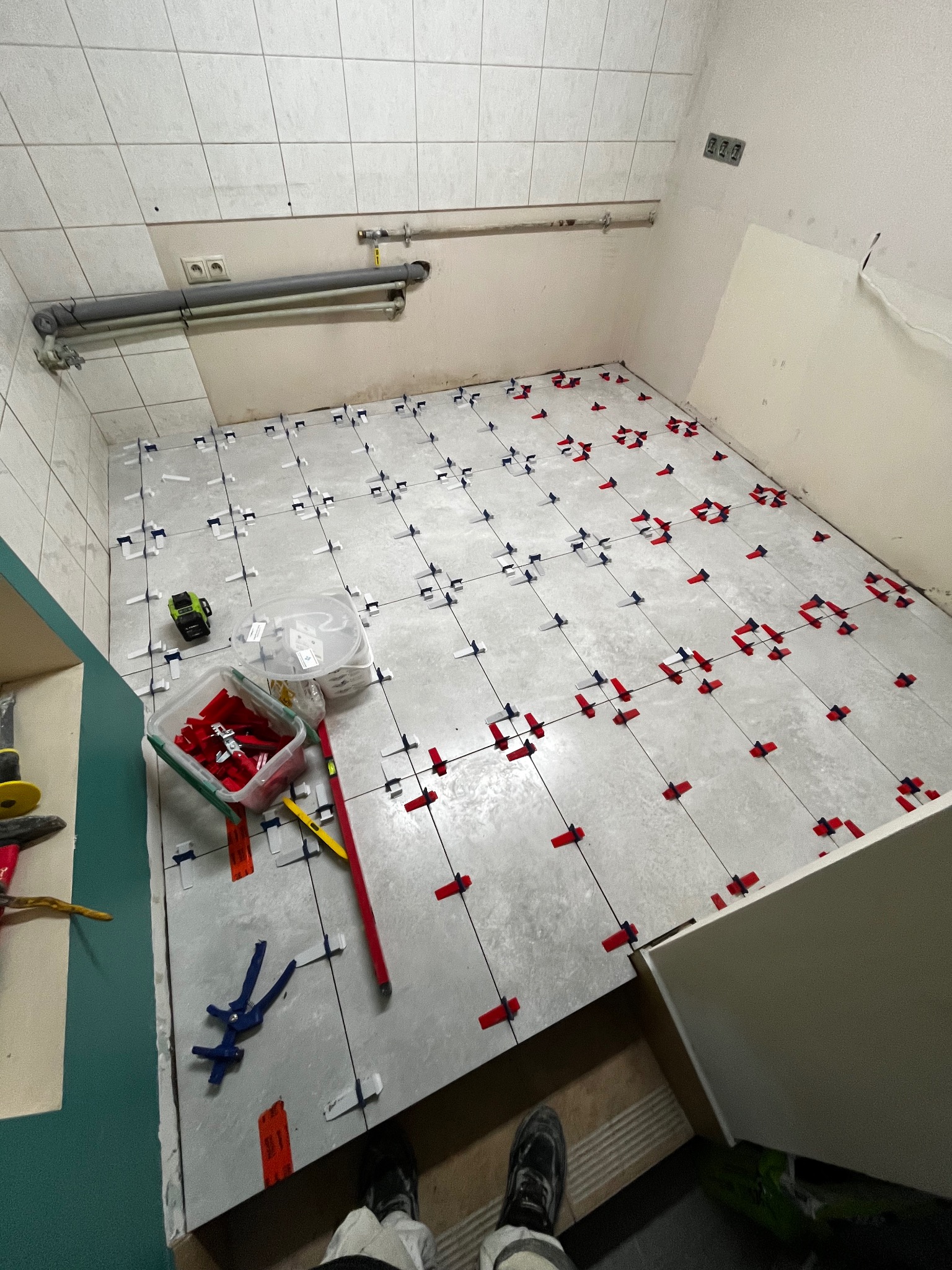 Układanie płytek podłogowych w łazience z użyciem systemu poziomowania. Widoczne narzędzia glazurnicze i fragment osoby w obuwiu roboczym. Szare płytki, czerwone i niebieskie kliny.