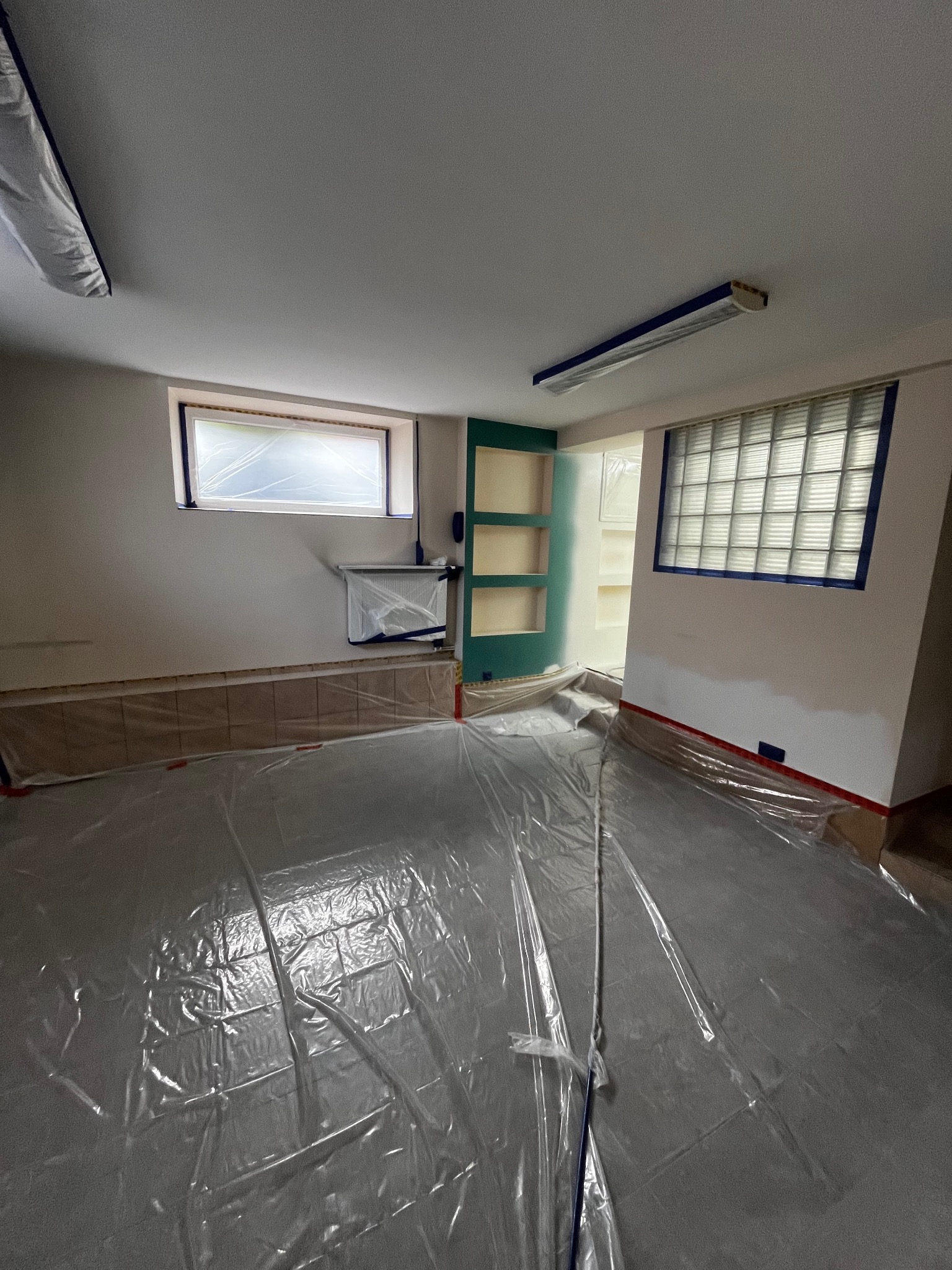 Przygotowane pomieszczenie do malowania: podłoga zabezpieczona folią, okna i listwy oklejone taśmą malarską. Widoczne półki wnękowe i grzejnik.