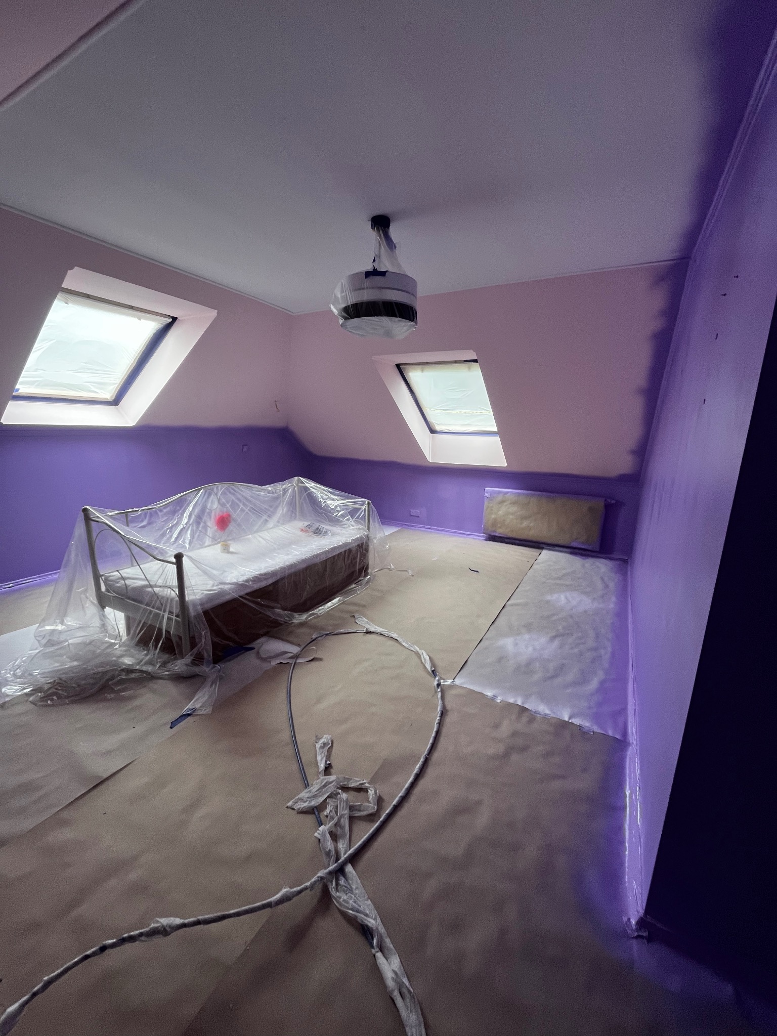 Pokój na poddaszu w trakcie malowania na fioletowo. Meble zabezpieczone folią, widoczne skosy i okna dachowe. Częściowo pomalowane ściany.