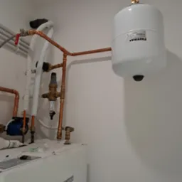 Biały zbiornik wyrównawczy Gitral połączony miedzianymi rurami z pompą ciepła Bosch, zawór bezpieczeństwa i manometr widoczne na instalacji.