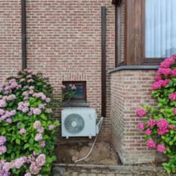 Biała jednostka zewnętrzna klimatyzacji przy ceglanym budynku, otoczona r&oacute;żowymi hortensjami. Widoczne okno i kostka brukowa.