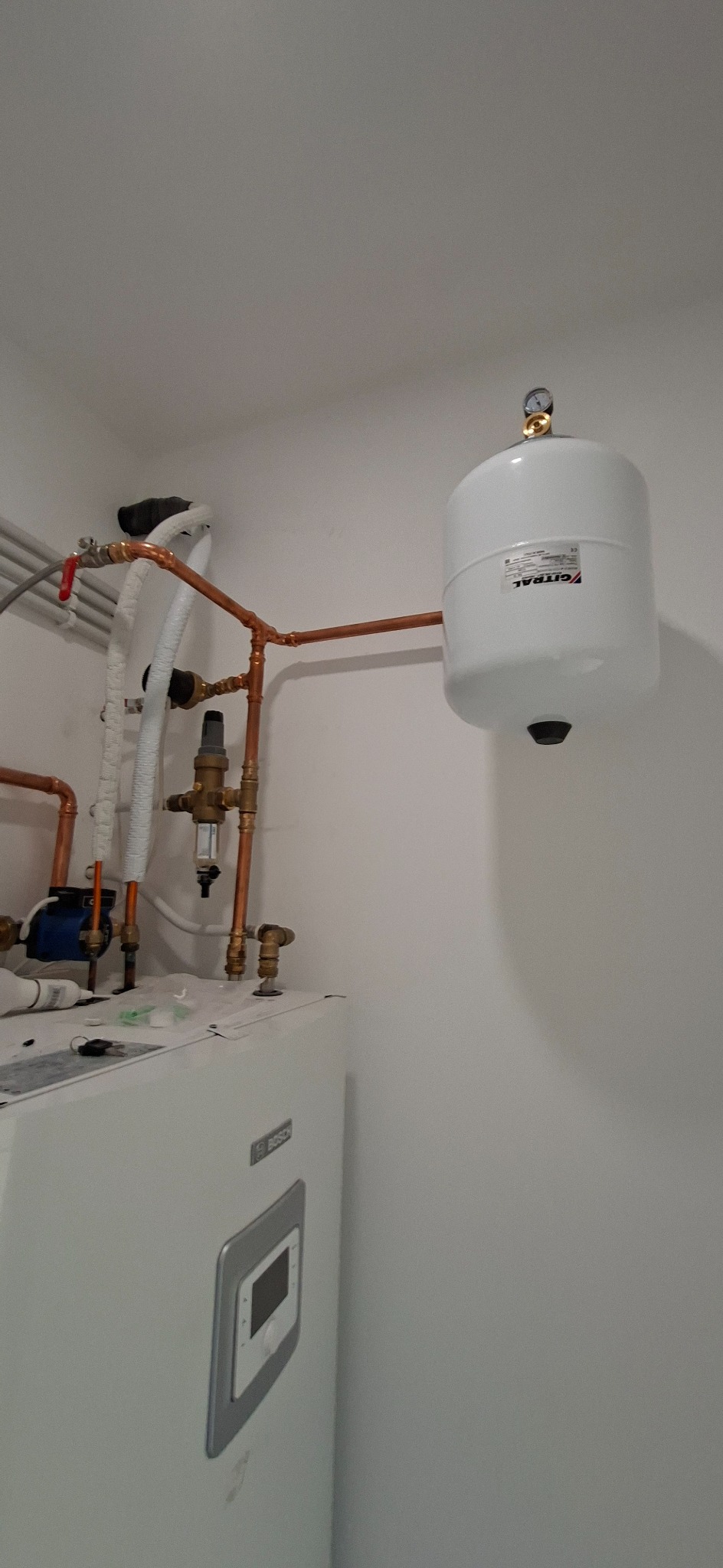 Biały zbiornik wyrównawczy Gitral połączony miedzianymi rurami z pompą ciepła Bosch, zawór bezpieczeństwa i manometr widoczne na instalacji.