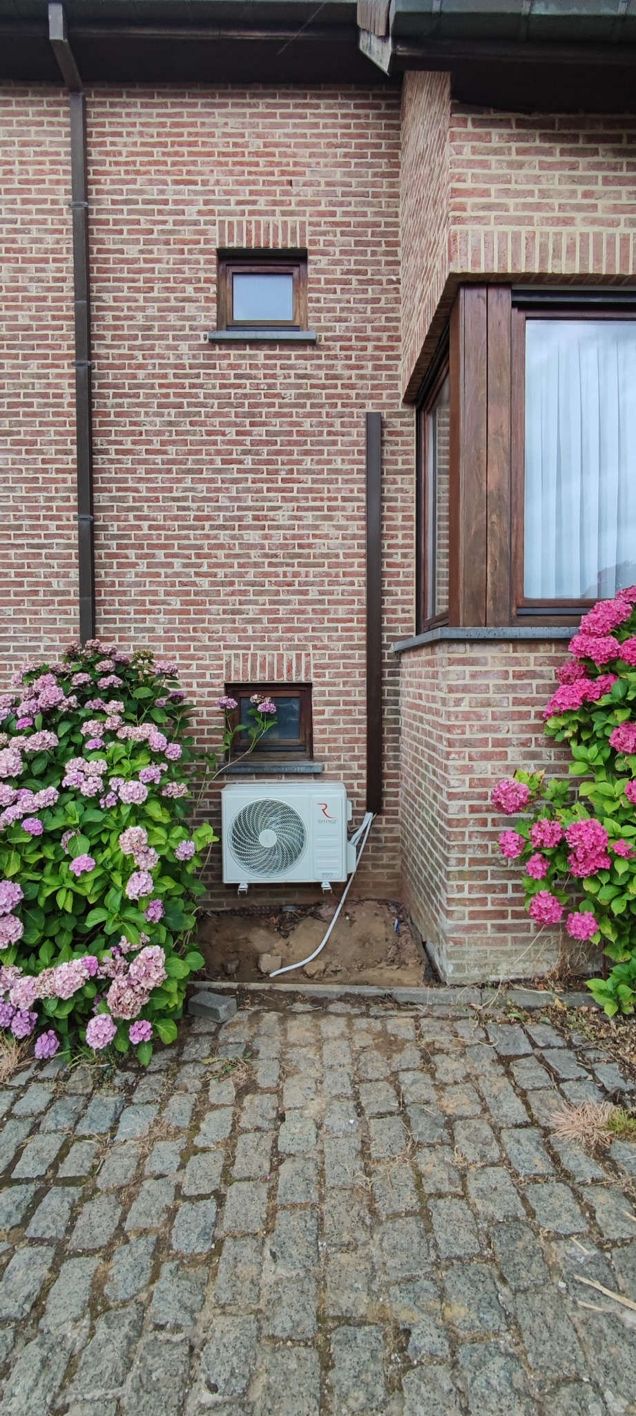 Biała jednostka zewnętrzna klimatyzacji przy ceglanym budynku, otoczona różowymi hortensjami. Widoczne okno i kostka brukowa.