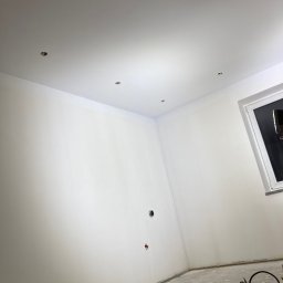 Zakład Handlowo Usługowy - Wnętrze pokoju po świeżym malowaniu na biało, widoczne punkty oświetleniowe w suficie, okno, oraz narzędzia malarskie i kabel przedłużający na podłodze.