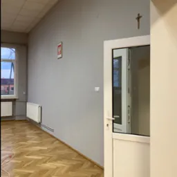 Wnętrze pomieszczenia po odświeżeniu: szare ściany, drewniana podłoga w jodełkę, białe drzwi z szybą i krzyż na ścianie.