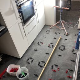 Zakład Handlowo Usługowy - Układanie ciemnoszarych płytek podłogowych w kuchni, widoczne krzyżyki dystansowe i poziomica, oświetlenie robocze na statywie.