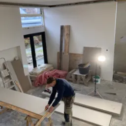 Pracownik w trakcie docinania płyty karton gips na drewnianym stojaku w pomieszczeniu w trakcie remontu, widoczne narzędzia, materiały budowlane i oświetlenie robocze.