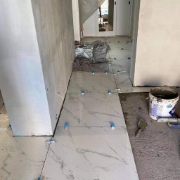 Zakład Handlowo Usługowy - Układanie jasnych płytek imitujących marmur na podłodze w korytarzu, z użyciem krzyżyków dystansowych, widoczny fragment ściany w stanie surowym i wiadro z zaprawą.