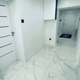 Wnętrze przedpokoju z białymi ścianami, marmurową podłogą, wbudowanymi punktowymi lampami w ścianie oraz białą szafką wiszącą z czarną obwódką.