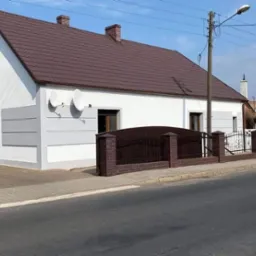 Biały, jednopiętrowy dom z brązowym dachem i dwoma antenami satelitarnymi na elewacji, widoczny od strony ulicy z ogrodzeniem z cegły i metalu.