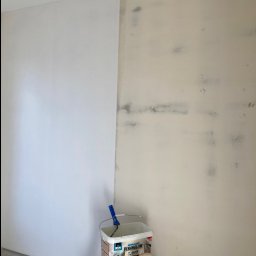 Zakład Handlowo Usługowy - Ściana w trakcie malowania: połowa pomalowana na biało, druga połowa w kolorze beżowym z widocznymi śladami po zabrudzeniach, wiadro z farbą i wałkiem na drabinie.