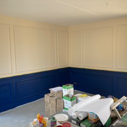 Zakład Handlowo Usługowy - Wnętrze pokoju z pomalowanymi na granatowo panelami ściennymi w dolnej części, oddzielonymi żółtą taśmą malarską od górnej, kremowej części ściany z dekoracyjnymi listwami. Na pierwszym planie...