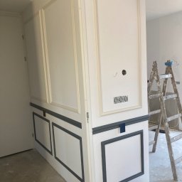 Zakład Handlowo Usługowy - Świeżo pomalowana ściana z dekoracyjnymi panelami w kolorze białym i szarym, widoczne gniazdka elektryczne, drabina malarska i wiadro z farbą, pomieszczenie w trakcie wykańczania.