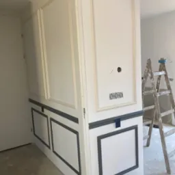 Świeżo pomalowana ściana z dekoracyjnymi panelami w kolorze białym i szarym, widoczne gniazdka elektryczne, drabina malarska i wiadro z farbą, pomieszczenie w trakcie wykańczania.