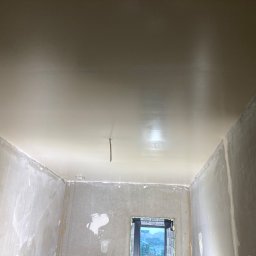 Zakład Handlowo Usługowy - Gładko wyszpachlowany sufit w pokoju z oknem, widoczne ściany przygotowane do dalszych prac wykończeniowych. Wystający kabel oświetleniowy.