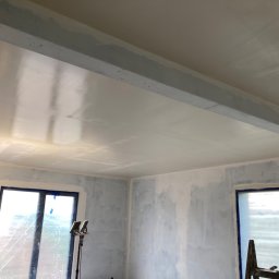 Zakład Handlowo Usługowy - Pokój w trakcie prac wykończeniowych: gładkie ściany i sufit po szpachlowaniu, okna zabezpieczone folią, widoczny pracownik z wałkiem.