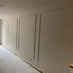 Pokój po remoncie: ściany z dekoracyjnymi listwami w odcieniach beżu, podłoga zabezpieczona folią malarską. Widoczny fragment sofy i okna.