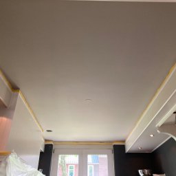 Zakład Handlowo Usługowy - Gładki, szary sufit po szpachlowaniu, zabezpieczony taśmą malarską przy ścianach w jasnej kuchni. Widoczne elementy zabudowy kuchennej i okno.