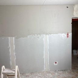 Zakład Handlowo Usługowy - Ściana w trakcie szpachlowania, widoczne łączenia płyt karton-gips, pracownik w kombinezonie nanosi gładź szpachlą. Fragment pokoju z widocznym wejściem do innego pomieszczenia.