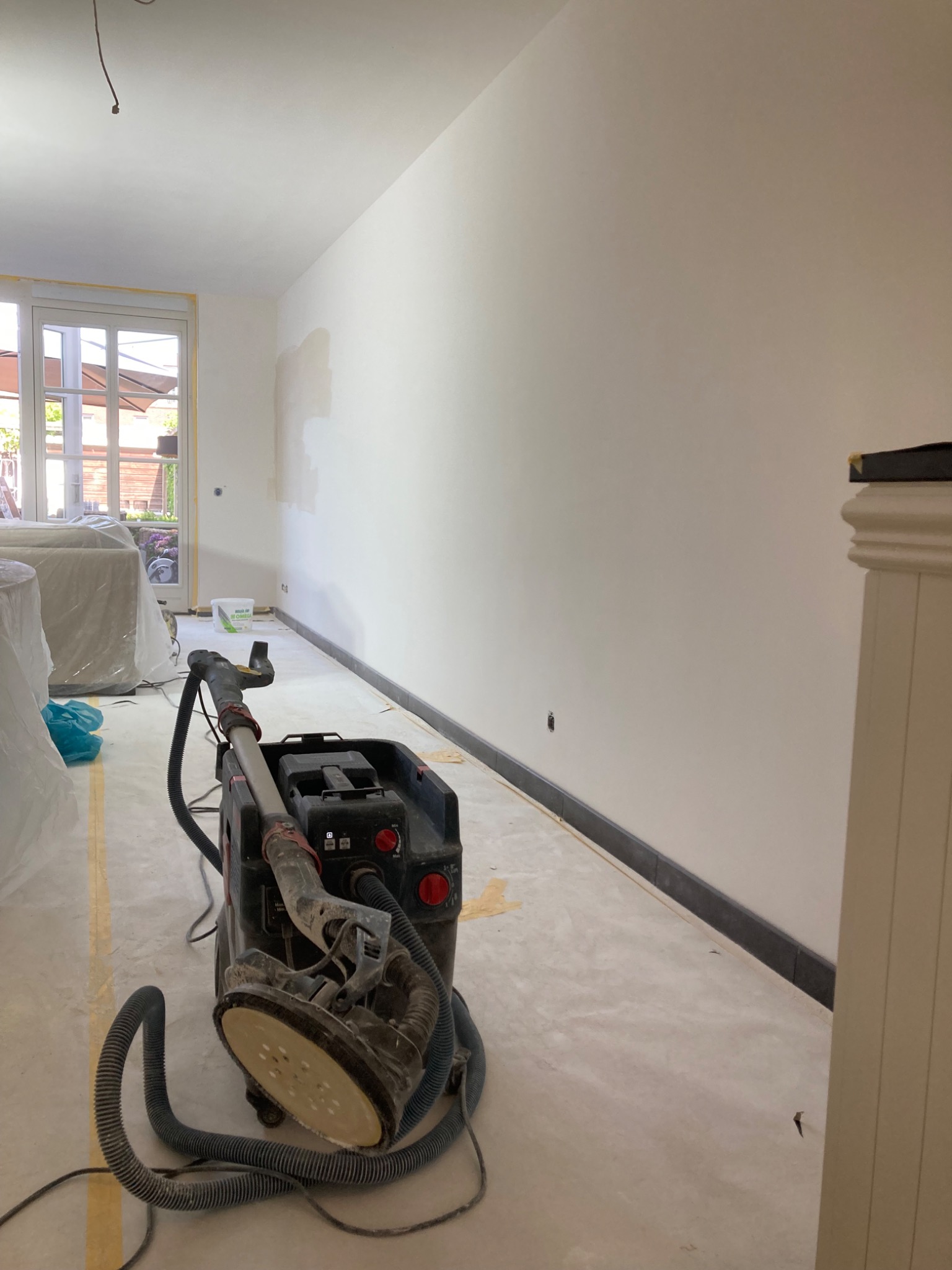 Szlifowanie ściany szlifierką teleskopową w pomieszczeniu z oknem i listwą przypodłogową. Przygotowanie do malowania, widoczne ślady po szpachlowaniu.