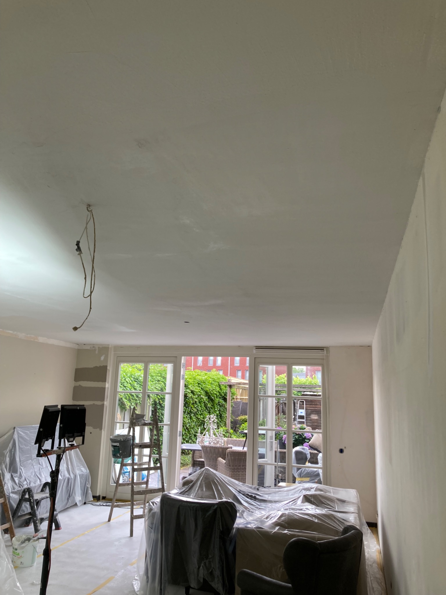 Wnętrze w trakcie prac: sufit po szpachlowaniu, zabezpieczone meble folią, drabina, widok na ogród przez przeszklone drzwi balkonowe, lampa budowlana.