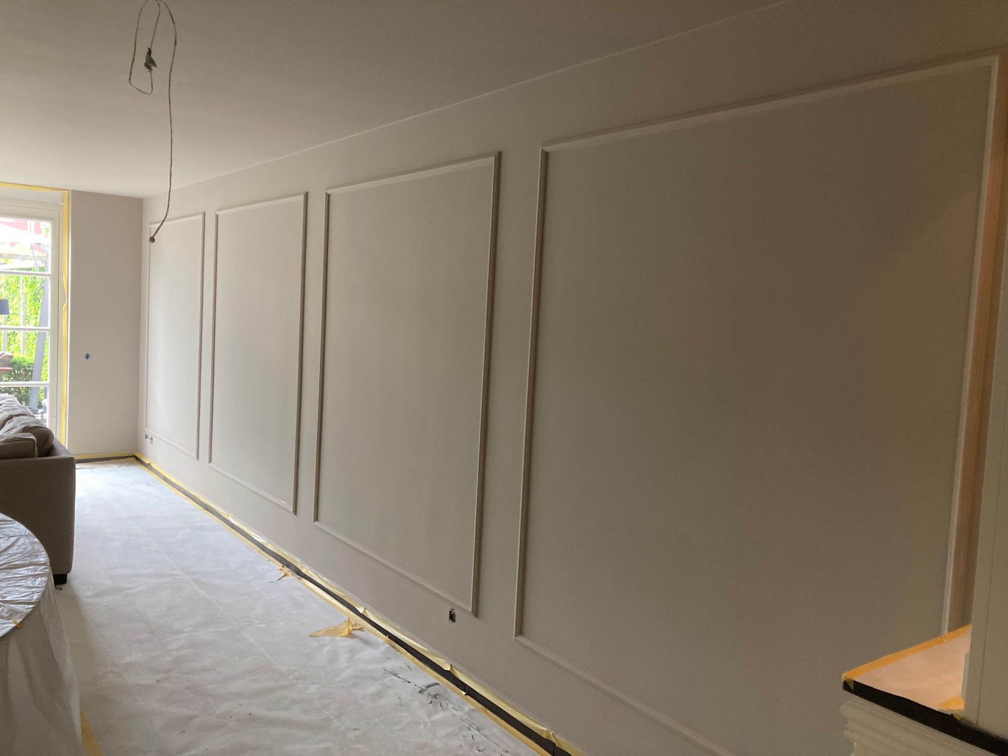 Pokój po remoncie: ściany z dekoracyjnymi listwami w odcieniach beżu, podłoga zabezpieczona folią malarską. Widoczny fragment sofy i okna.