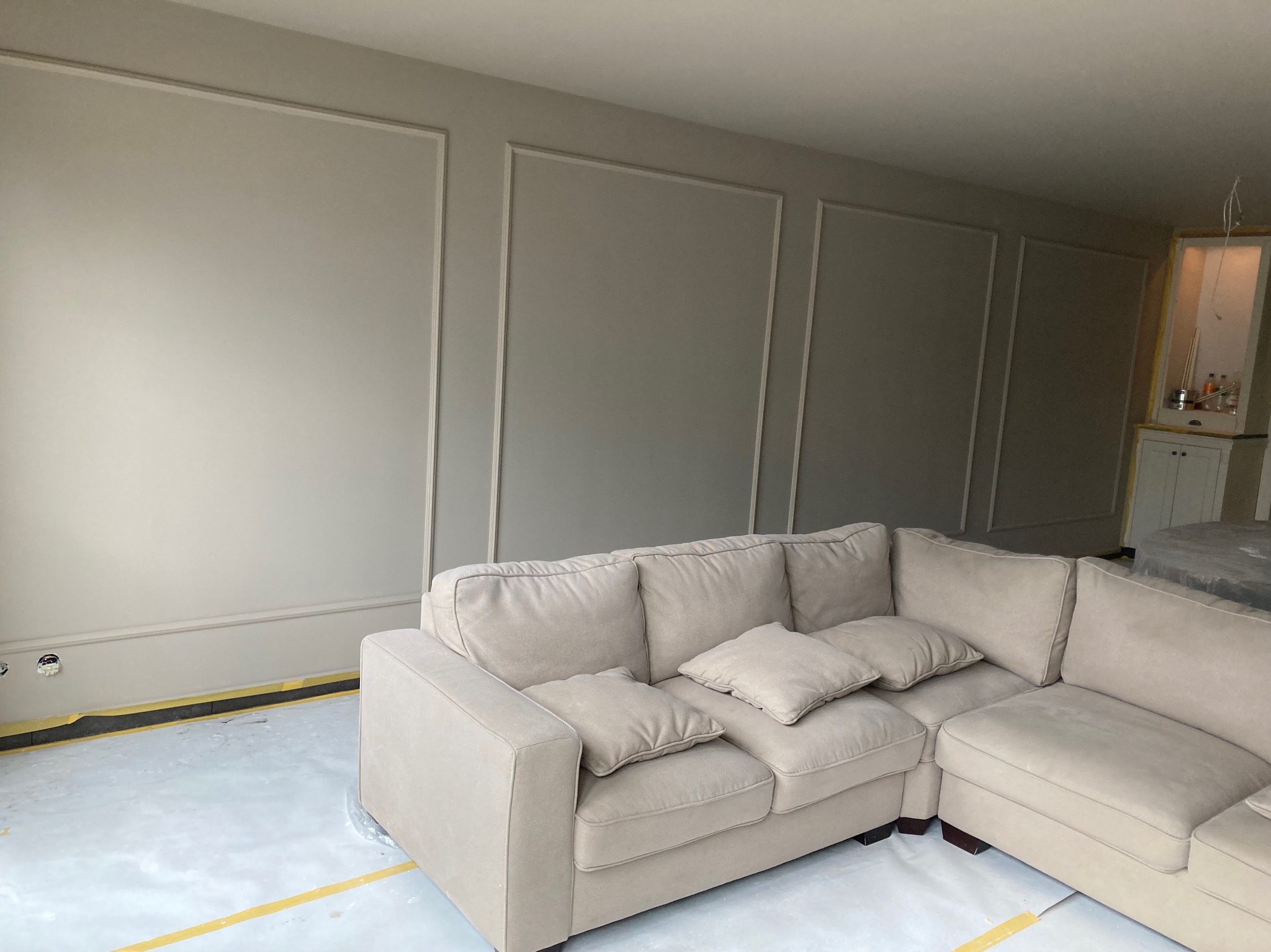 Jasnoszara ściana z dekoracyjnymi listwami w prostokątnych ramach nad beżową sofą. Podłoga zabezpieczona folią i taśmą malarską, widoczne gniazdka elektryczne.