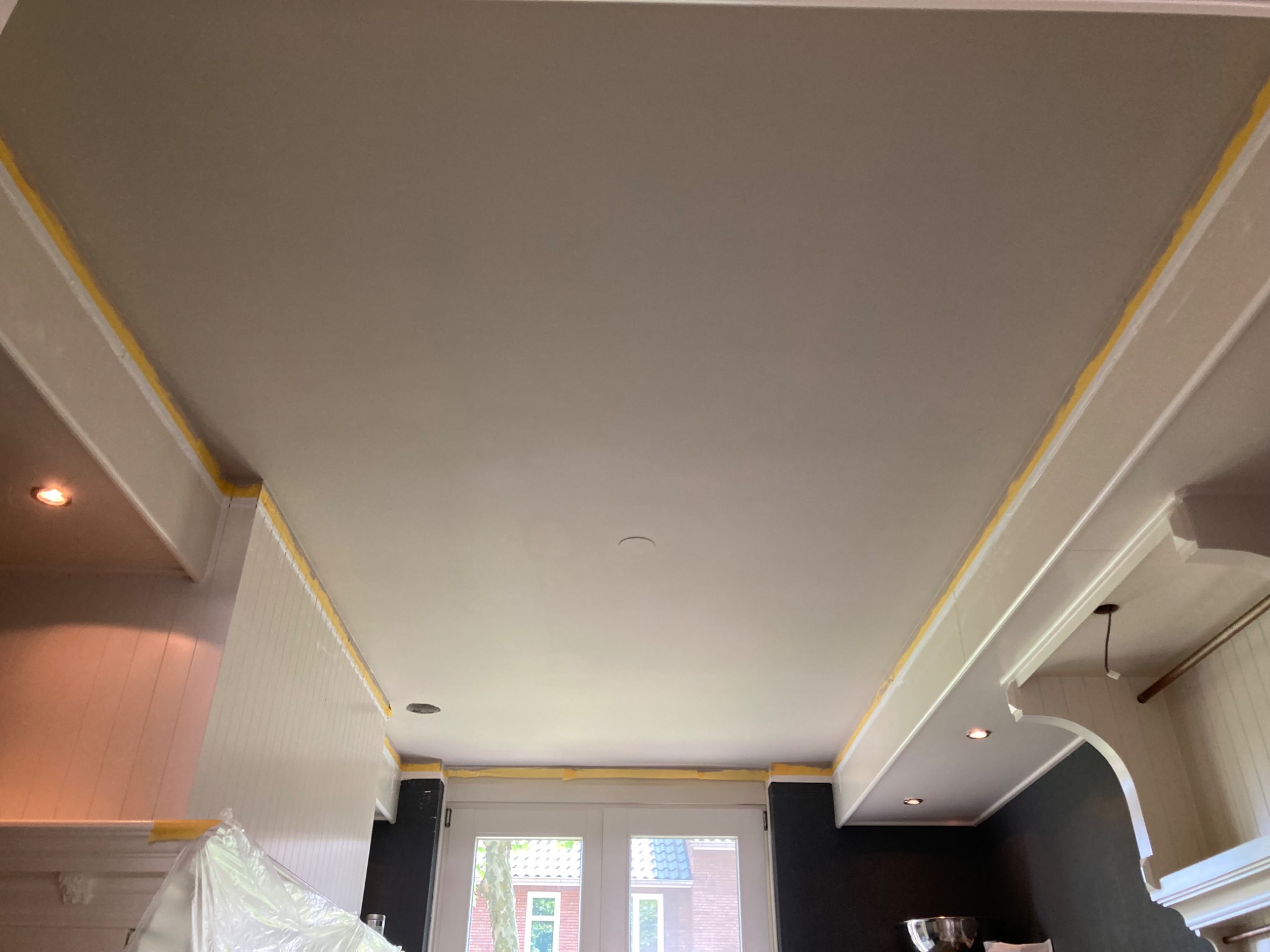 Gładki, szary sufit po szpachlowaniu, zabezpieczony taśmą malarską przy ścianach w jasnej kuchni. Widoczne elementy zabudowy kuchennej i okno.