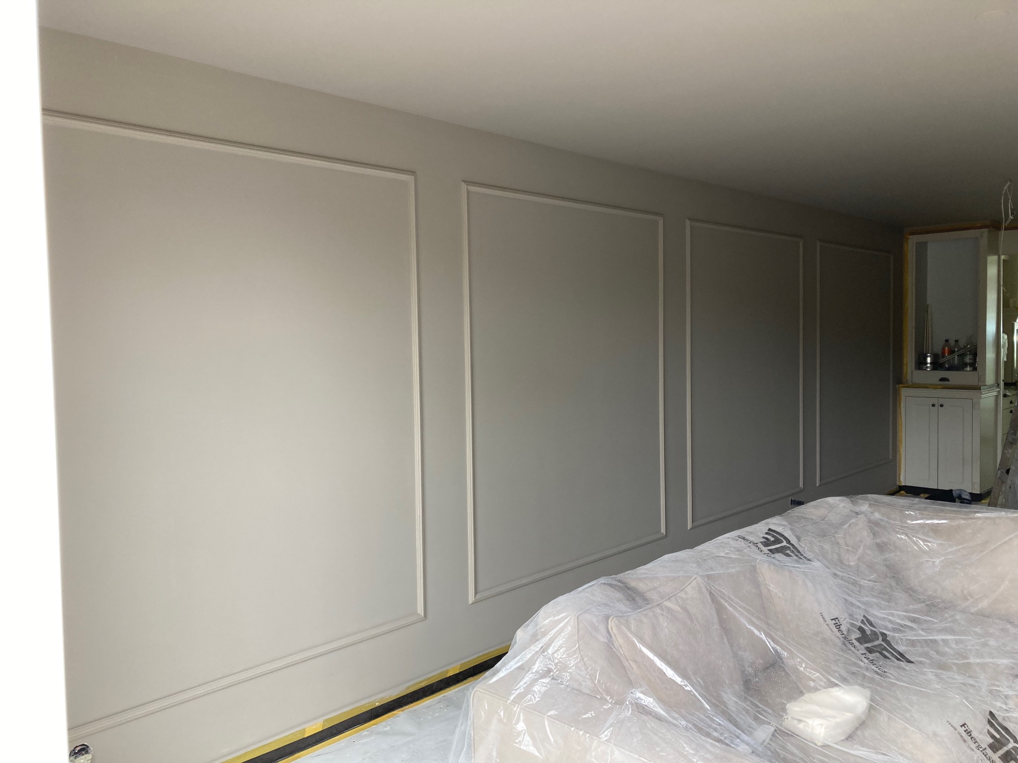 Szare ściany z dekoracyjnymi listwami w ramkach, zabezpieczone meble folią malarską. Widok na wnętrze w trakcie prac wykończeniowych, z elementami zabudowy w tle.