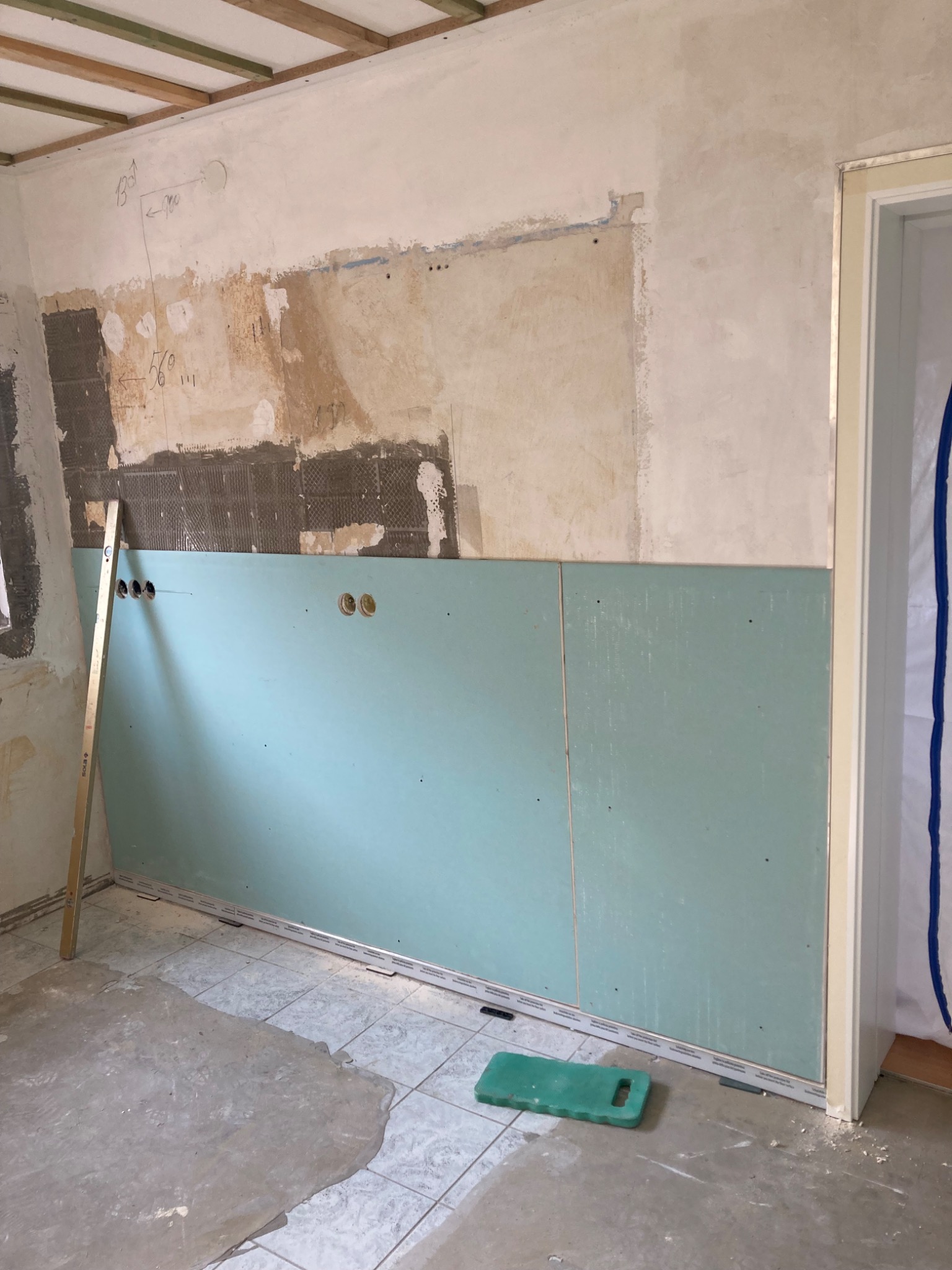 Montaż płyty karton-gips w trakcie remontu: zielona płyta częściowo zamontowana na ścianie z widocznymi otworami na gniazdka, poziomica oparta o ścianę, surowy stan podłogi.