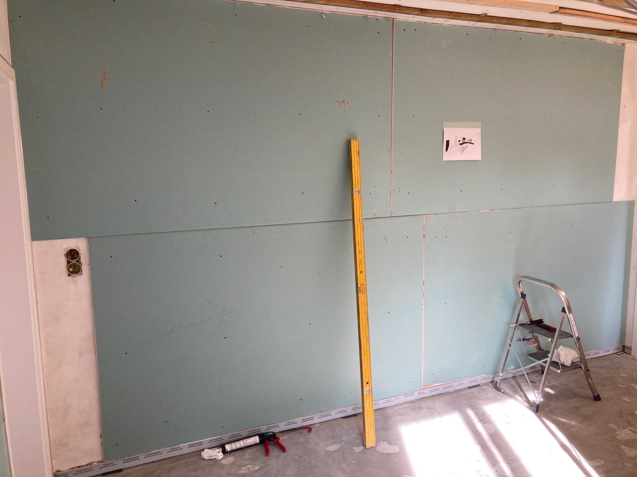 Montaż płyt karton-gips na ścianie z widoczną poziomicą, drabinką i elementami wykończeniowymi. Widoczne gniazdka elektryczne i plan instalacji na ścianie.