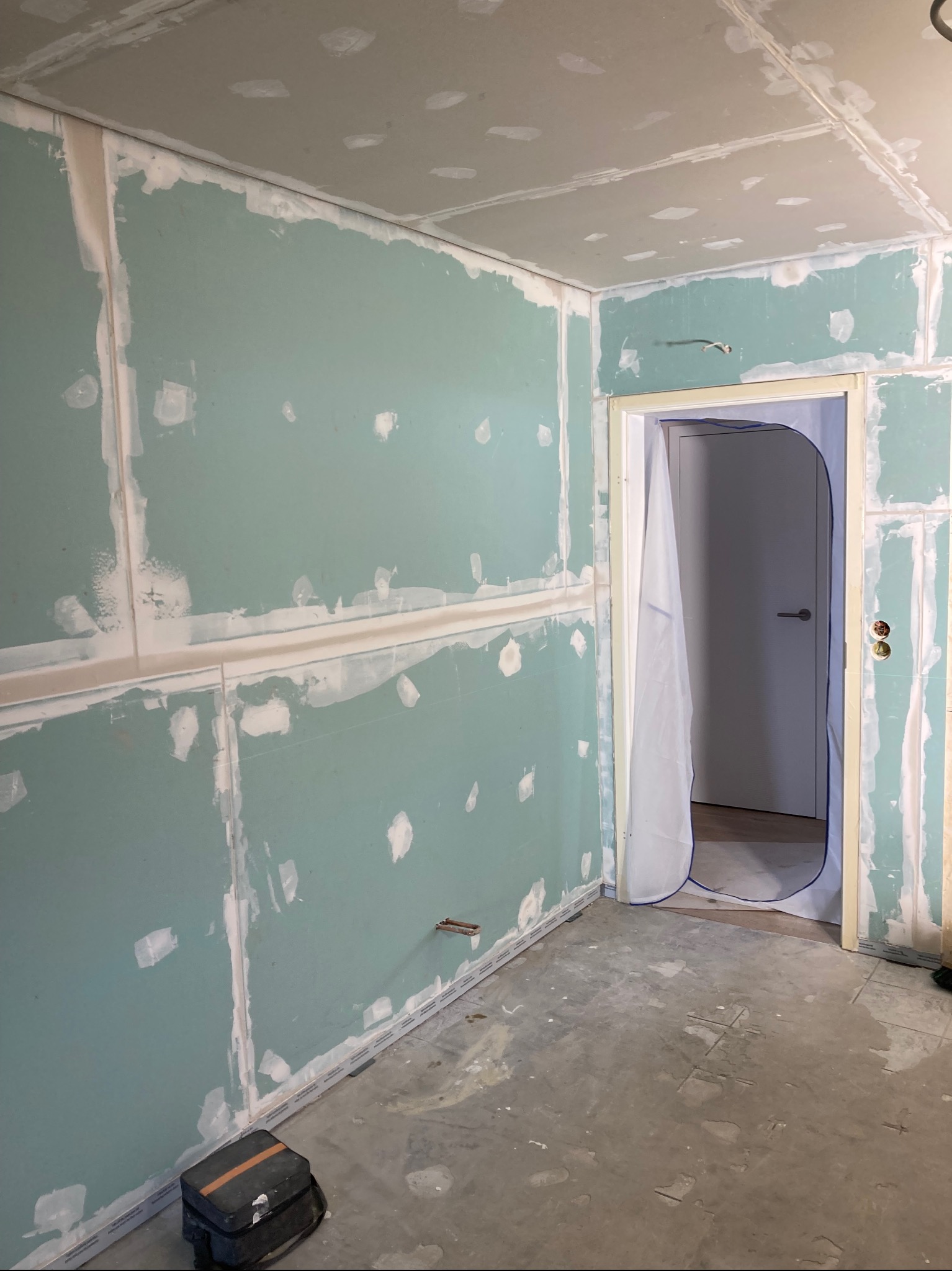 Pokój w trakcie prac: płyty karton-gips na ścianach i suficie, spoiny zaszpachlowane, zabezpieczone drzwi folią. Widoczny fragment podłogi i narzędzia.