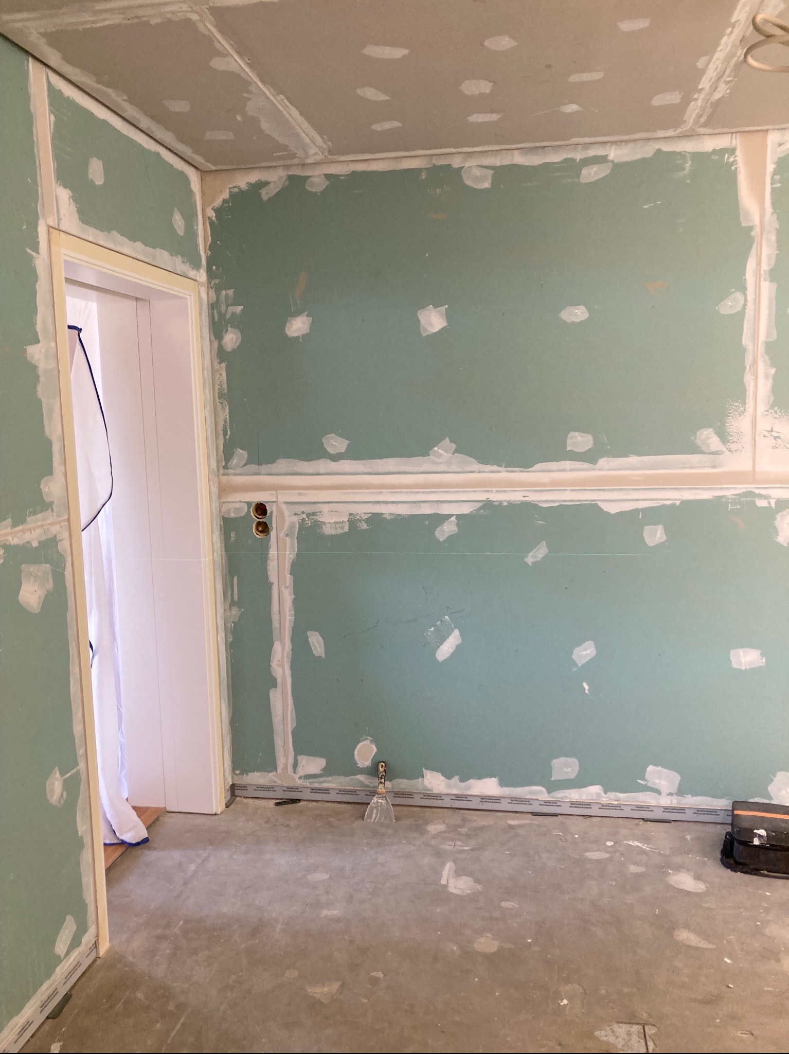 Wnętrze w trakcie prac: ściany wyłożone płytami kartonowo-gipsowymi, widoczne łączenia i miejsca po wkrętach przygotowane do szpachlowania. Otwór drzwiowy z ościeżnicą.